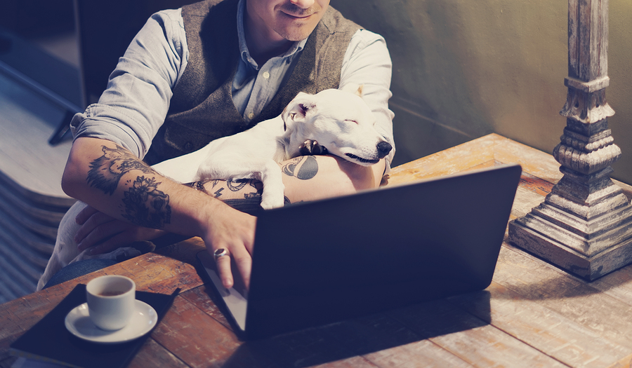 Smiling tattooed man in eyeglasses working at home on laptop while sitting at the wooden table with cute dog sleeping on his hands.Adult guy using modern computer for surfing web.Horizontal