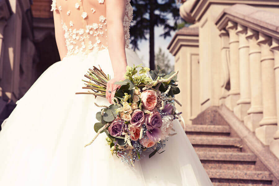 Bride stood on steps wearing white lace and tulle wedding dress holding bouquet of flowers