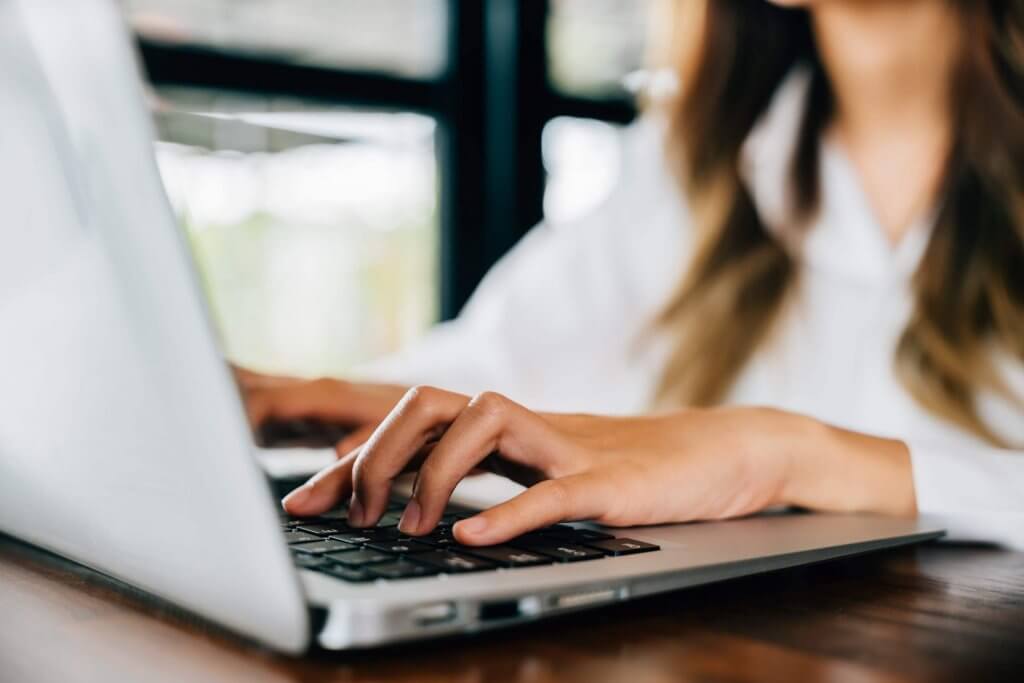 A woman with long brown hair, wearing a white shirt, typing on a silver laptop.